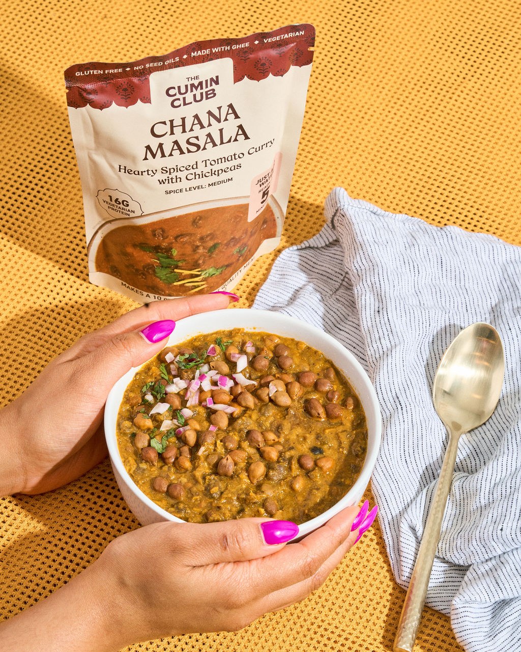 Hands holding a bowl of Chana Masala beside the pack on a yellow backdrop